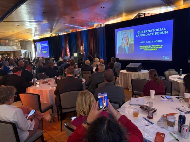 A full ballroom of attendees at round tables facing screens that show Governor Katie Hobbs for the Gubernatorial Candidate Forum.