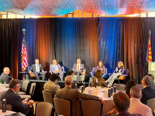 Five panelists seated on stage between the American and Arizona flags, with attendees watching from the tables in front.