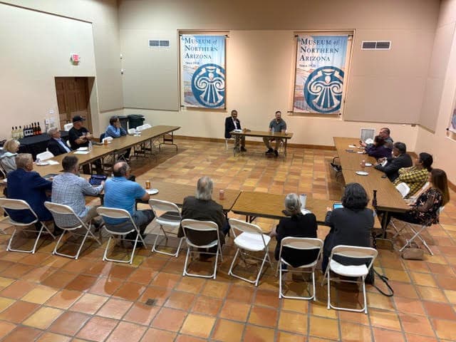 Members and candidates seated at folding tables in a rectangular arrangement inside the Museum of Northern Arizona, with two candidates at the center taking questions from the group.