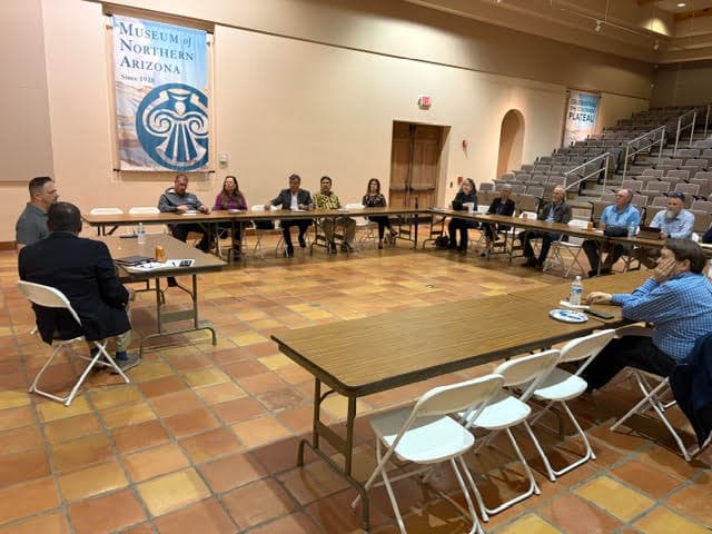 A wider view of the same room showing the full group around the tables, with the museum's tiered auditorium seating in the background.